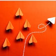 White paper plane is flying to the right on orange colored background where a group of orange colored paper planes are flying straight up.