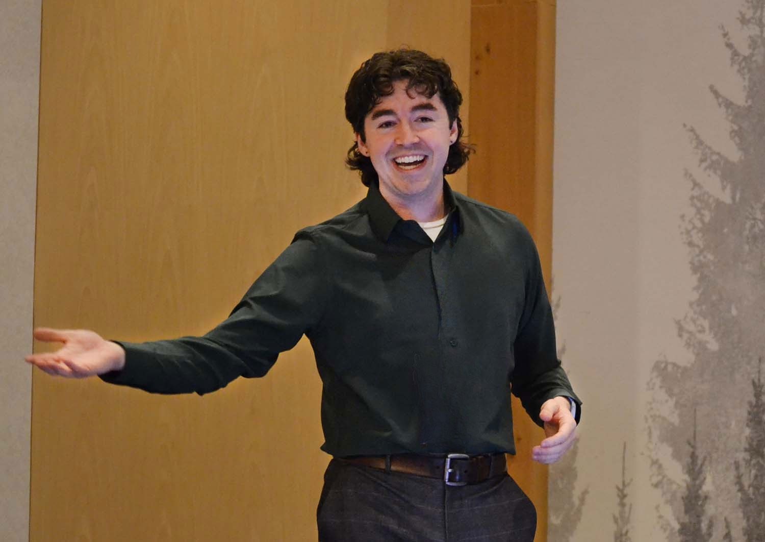 Young man with dark curly hair smiling while gesturing with open arms in a bright indoor space