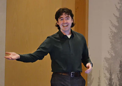 Young man with dark curly hair smiling while gesturing with open arms in a bright indoor space