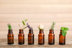 Six amber glass bottles with fresh herbs and flowers including rosemary, lavender, and mint arranged on wooden surface