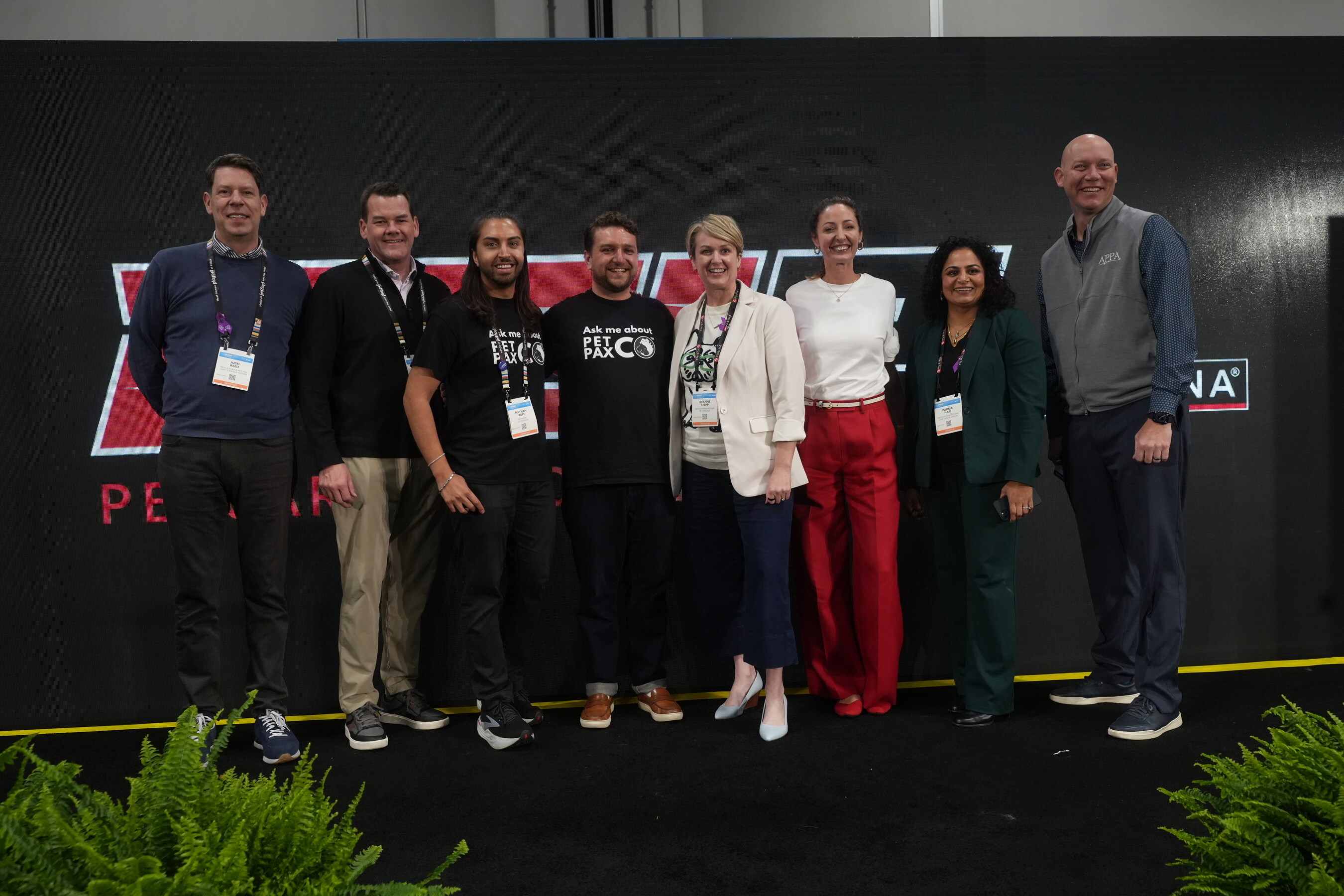 Eight professionals wearing conference badges pose together at a branded event backdrop