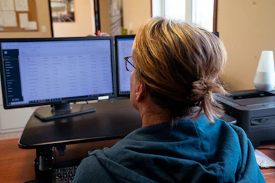 Woman working at office desk with dual computer monitors displaying spreadsheet data