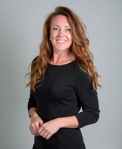 Professional headshot of a smiling woman with long reddish-brown wavy hair wearing a black three-quarter sleeve top
