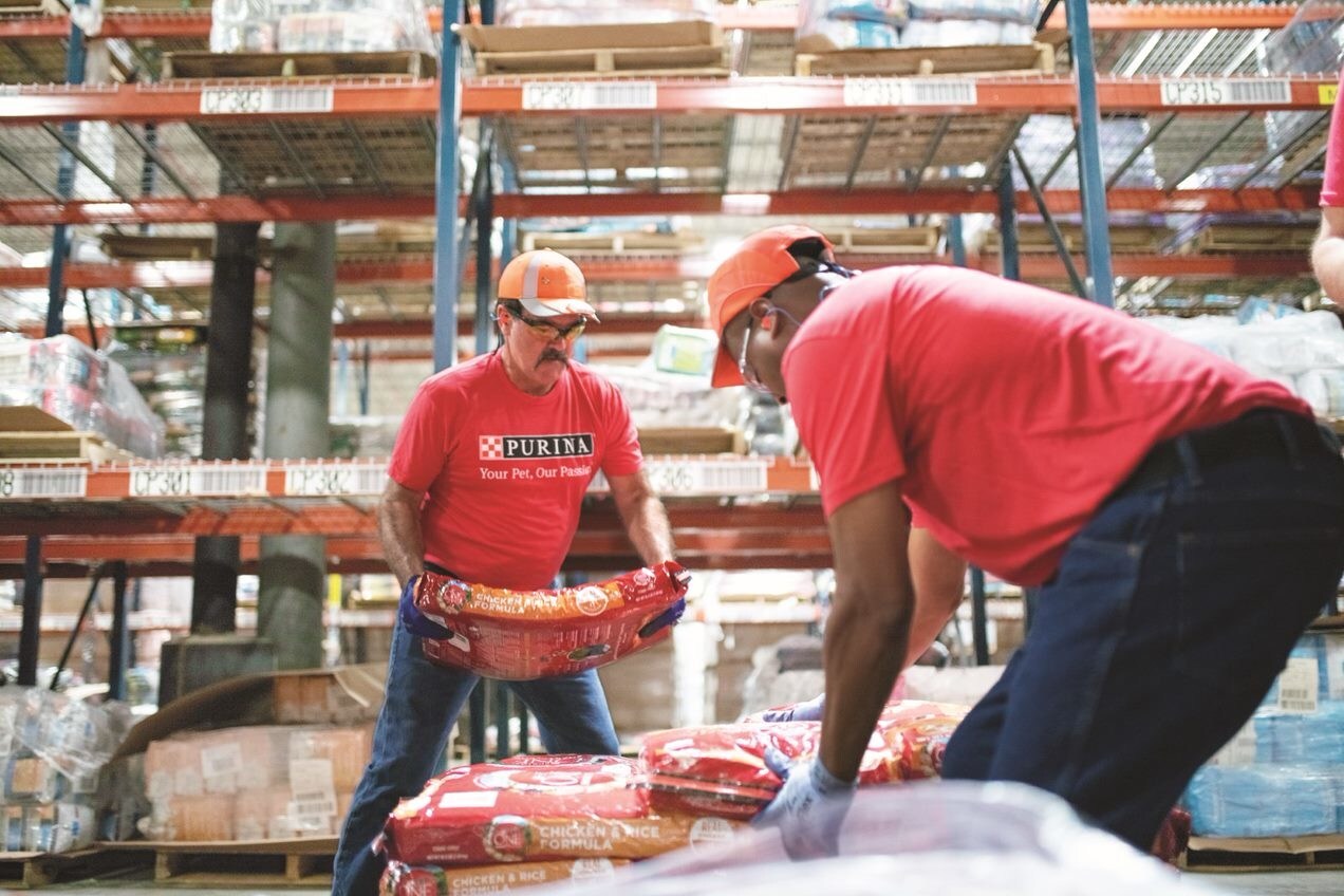 Warehouse workers in red Purina shirts handling pet food packages on pallets in industrial storage facility