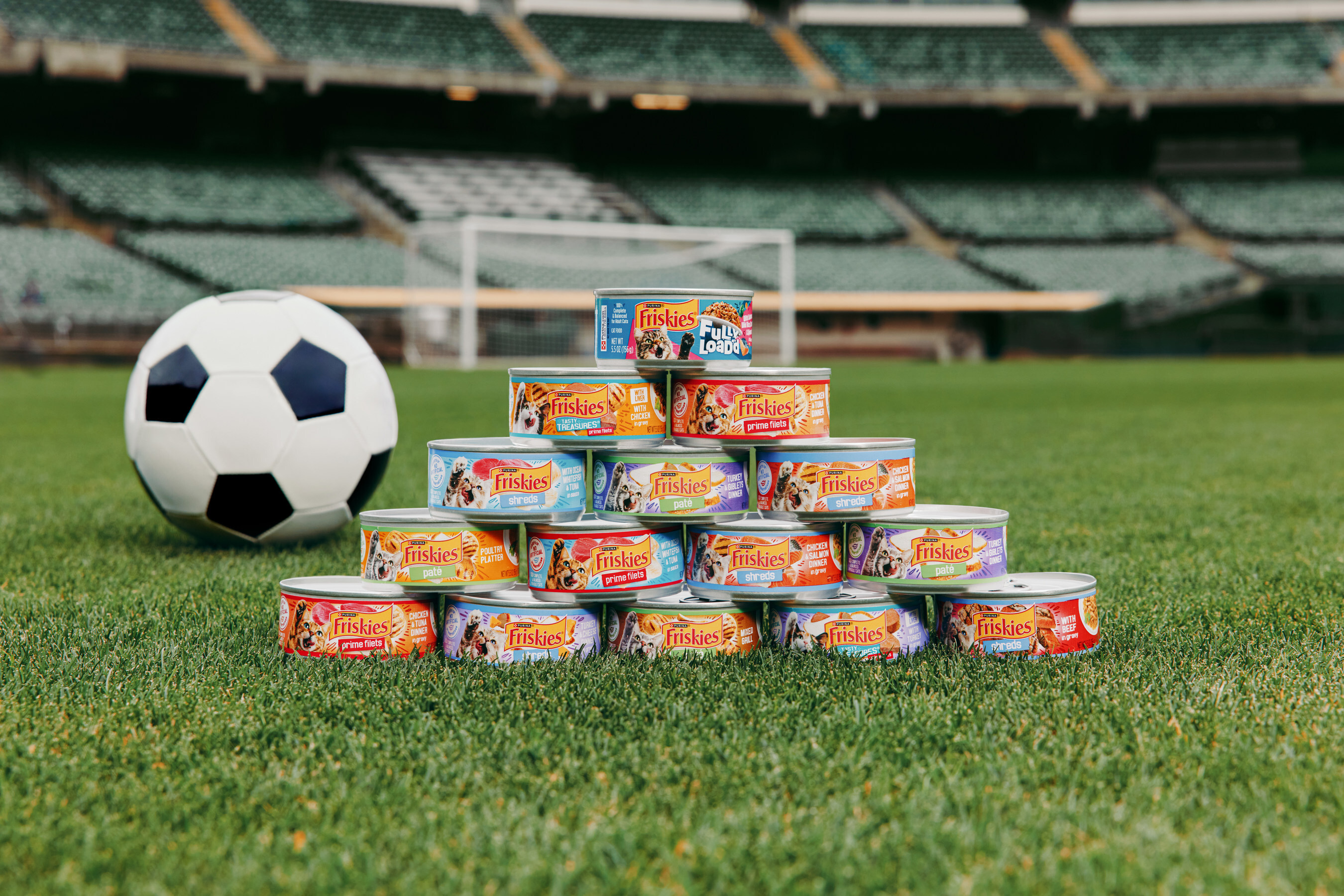 Soccer ball and pyramid stack of Friskies pet food cans on stadium grass field