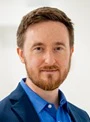 Professional headshot of a man with beard wearing blue shirt against neutral background