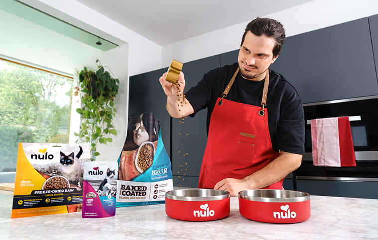 Man in red apron holding Nulo pet food treat in modern kitchen with product bowls and packaging
