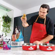 Man in red apron holding Nulo pet food treat in modern kitchen with product bowls and packaging