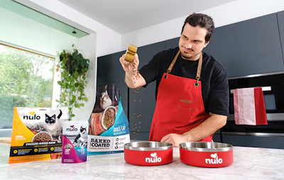 Man in red apron holding Nulo pet food treat in modern kitchen with product bowls and packaging