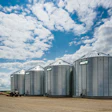 Row of silver grain storage silos with AGI branding under blue cloudy sky on agricultural farm