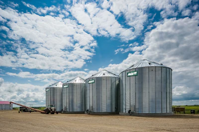 Row of silver grain storage silos with AGI branding under blue cloudy sky on agricultural farm