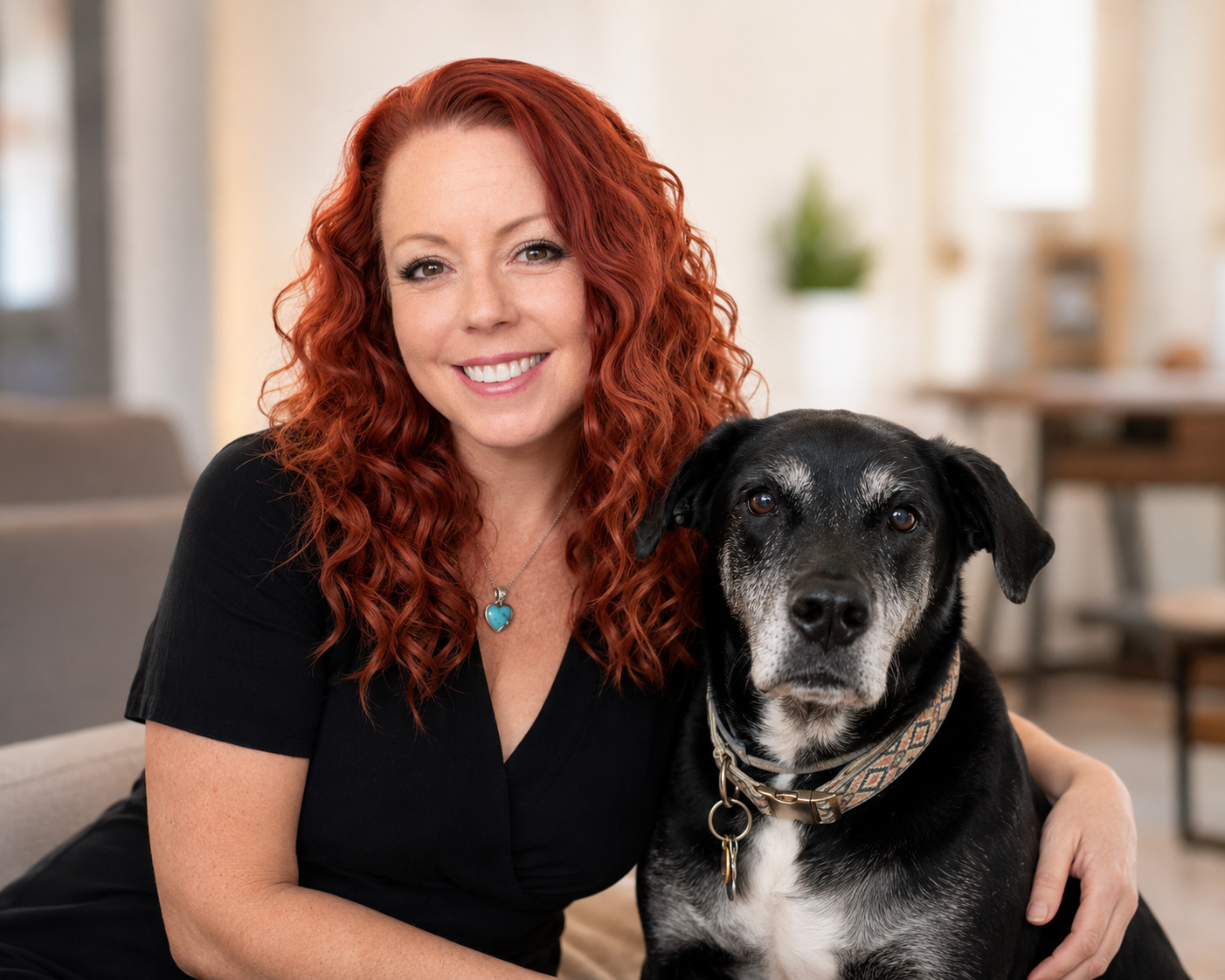 Woman with red curly hair smiling with senior black dog wearing collar in modern home interior