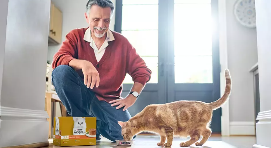 Middle-aged man crouching beside an orange tabby cat with pet food box in bright hallway