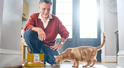Middle-aged man crouching beside an orange tabby cat with pet food box in bright hallway