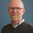 Professional headshot of a bearded man wearing glasses and a dark sweater over a checkered shirt against a blue background
