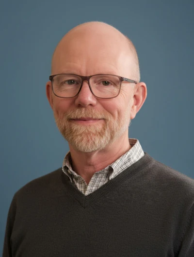 Professional headshot of a bearded man wearing glasses and a dark sweater over a checkered shirt against a blue background