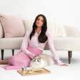 Woman in pink outfit sitting with white cat on cream sofa, pet food products on wooden tray
