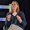 Professional woman speaking at conference stage with blue striped backdrop, wearing navy cardigan and gesturing expressively