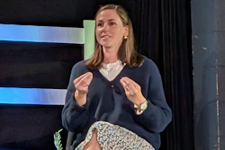 Professional woman speaking at conference stage with blue striped backdrop, wearing navy cardigan and gesturing expressively
