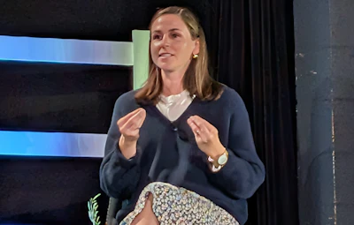 Professional woman speaking at conference stage with blue striped backdrop, wearing navy cardigan and gesturing expressively
