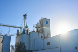 Industrial grain elevator facility with metal silos, conveyor systems, and white processing building under clear blue sky