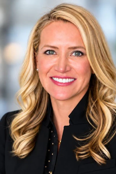 Professional headshot of a blonde woman smiling, wearing a black blazer against a blurred background