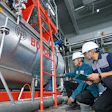Two technicians in safety gear inspect a red and silver industrial boiler system in a factory warehouse