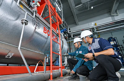 Two technicians in safety gear inspect a red and silver industrial boiler system in a factory warehouse