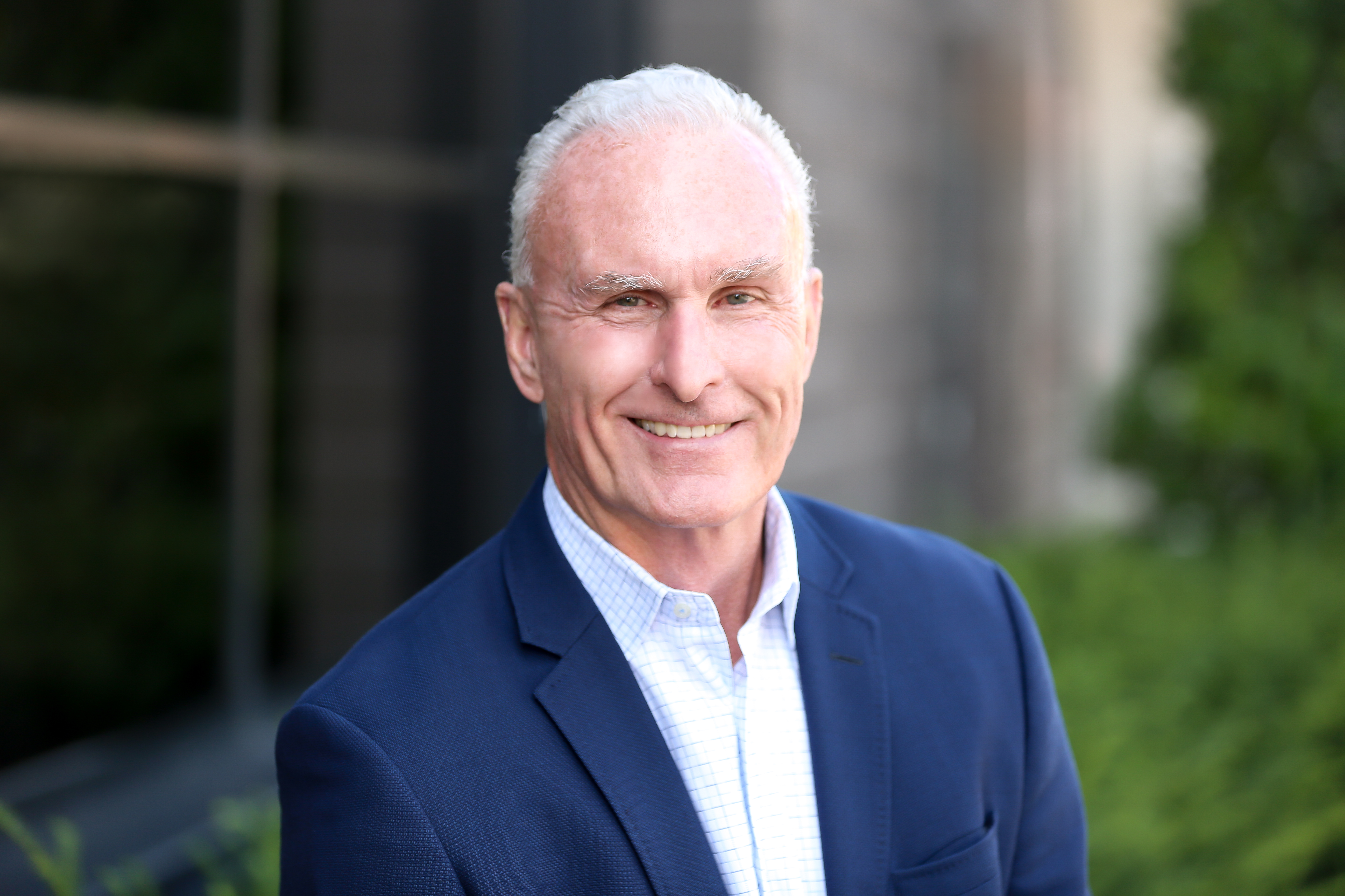 Professional headshot of a smiling man in navy blazer and light blue shirt against blurred architectural background