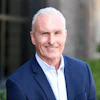 Professional headshot of a smiling man in navy blazer and light blue shirt against blurred architectural background