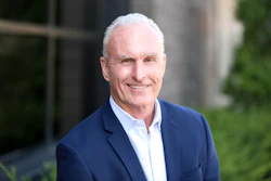 Professional headshot of a smiling man in navy blazer and light blue shirt against blurred architectural background