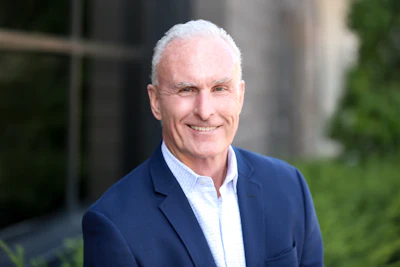 Professional headshot of a smiling man in navy blazer and light blue shirt against blurred architectural background