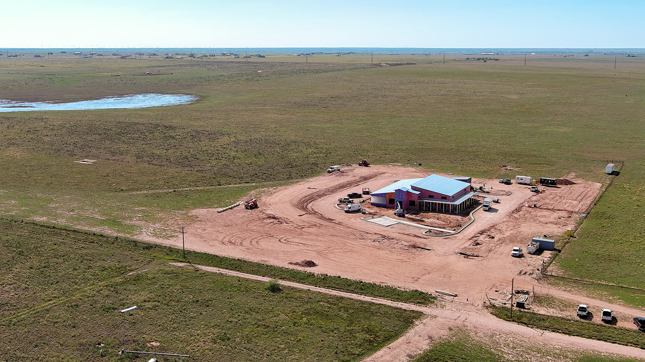 Aerial view of rural construction site with red barn and blue roof surrounded by green fields and dirt roads
