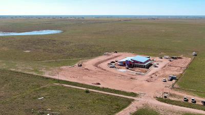 Aerial view of rural construction site with red barn and blue roof surrounded by green fields and dirt roads