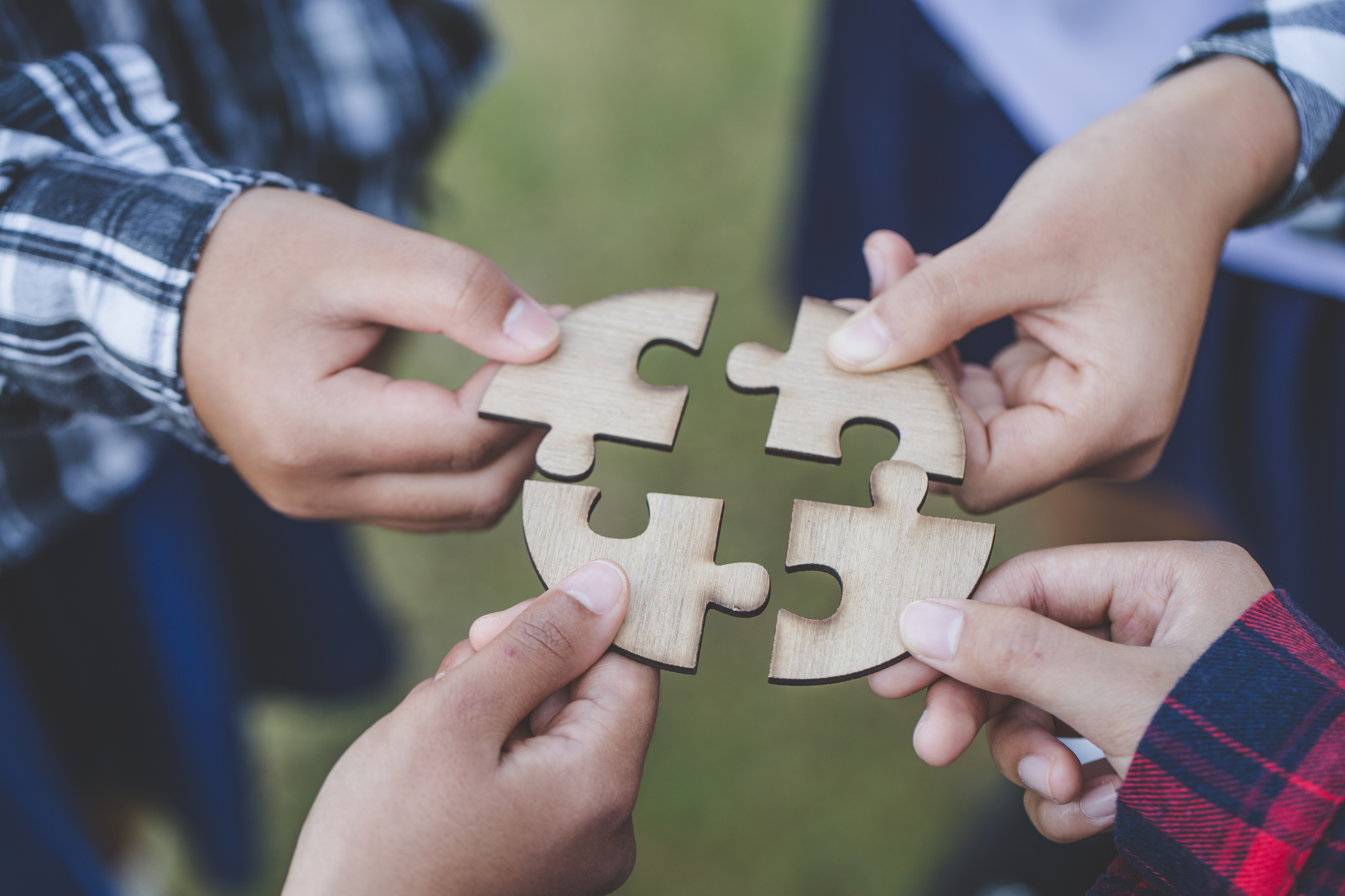 Four hands holding interlocking wooden puzzle pieces together outdoors, symbolizing teamwork and collaboration
