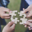 Four hands holding interlocking wooden puzzle pieces together outdoors, symbolizing teamwork and collaboration