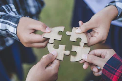 Four hands holding interlocking wooden puzzle pieces together outdoors, symbolizing teamwork and collaboration