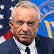 Professional headshot of an older man in dark pinstriped suit and tie, with American flag and official government seal in background