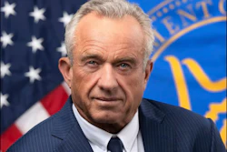 Professional headshot of an older man in dark pinstriped suit and tie, with American flag and official government seal in background