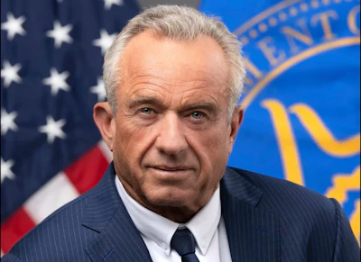 Professional headshot of an older man in dark pinstriped suit and tie, with American flag and official government seal in background