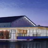 Modern waterfront building with pitched roof, wood and metal cladding, illuminated glass facade at dusk
