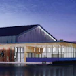 Modern waterfront building with pitched roof, wood and metal cladding, illuminated glass facade at dusk