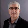 Professional headshot of a man with gray hair wearing glasses and dark blazer over gray shirt