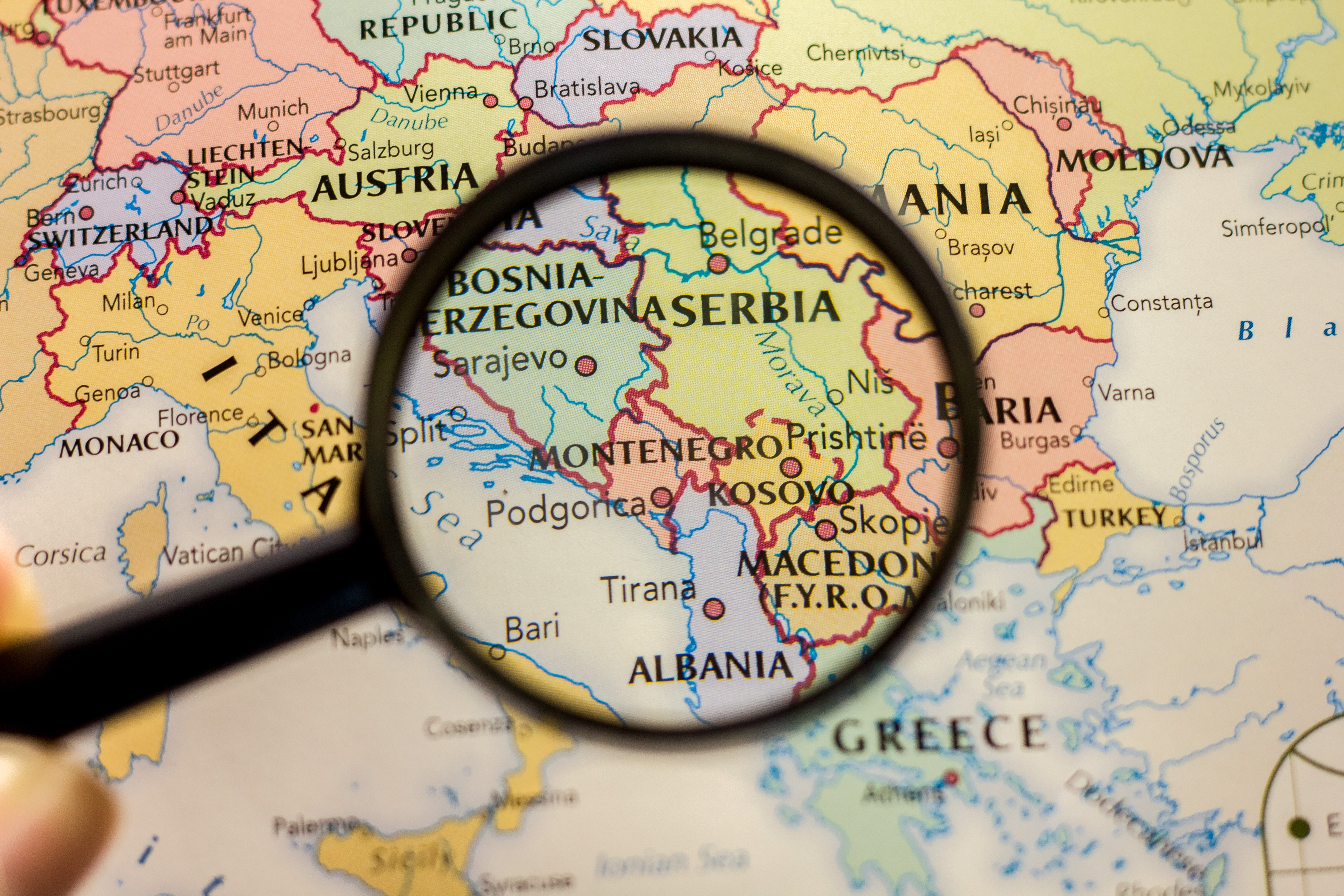 Magnifying glass examining a map of the Balkans region showing Serbia, Bosnia, Kosovo, and surrounding countries