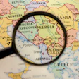 Magnifying glass examining a map of the Balkans region showing Serbia, Bosnia, Kosovo, and surrounding countries