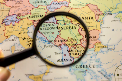 Magnifying glass examining a map of the Balkans region showing Serbia, Bosnia, Kosovo, and surrounding countries